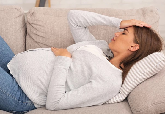 Schwangere Frau liegt auf dem Sofa und hält sich die Stirn und Bauch. 