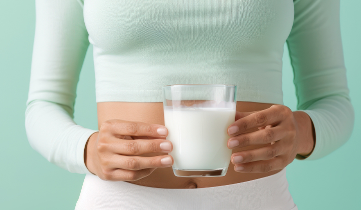 Une femme avec un haut turquoise et un pantalon blanc tient un verre de lait des deux mains.