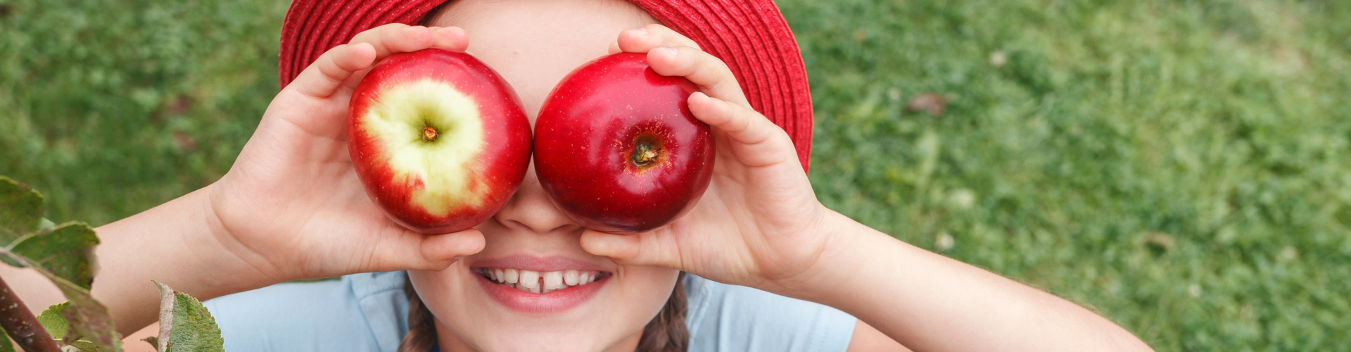 Une fillette avec un T-shirt bleu clair, une petite robe en jean et un chapeau de soleil rouge, les cheveux tressés à gauche et à droite, joue dans le jardin sous un pommier et tient deux pommes rouges devant ses yeux.