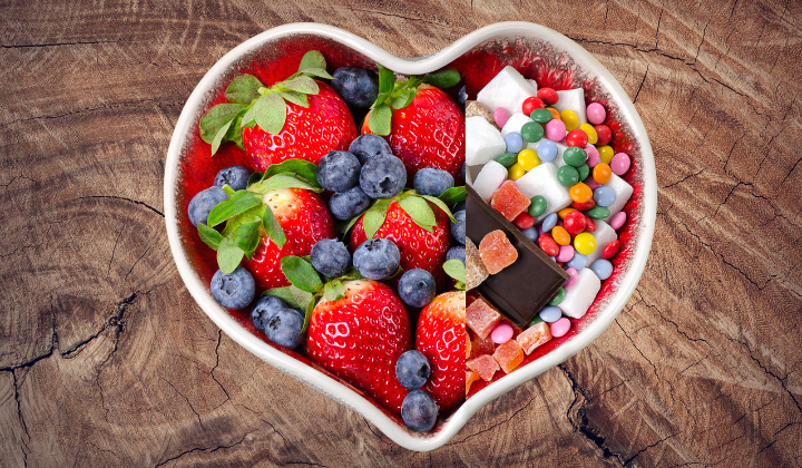 Weisse Herzschale. Die eine Haelfte ist gefuellt mit Erdbeeren, Blaubeeren und die andere Haelfte mit Suessigkeiten. Die Schale steht auf einem Holztisch. Alles aus Vogelperspektive.